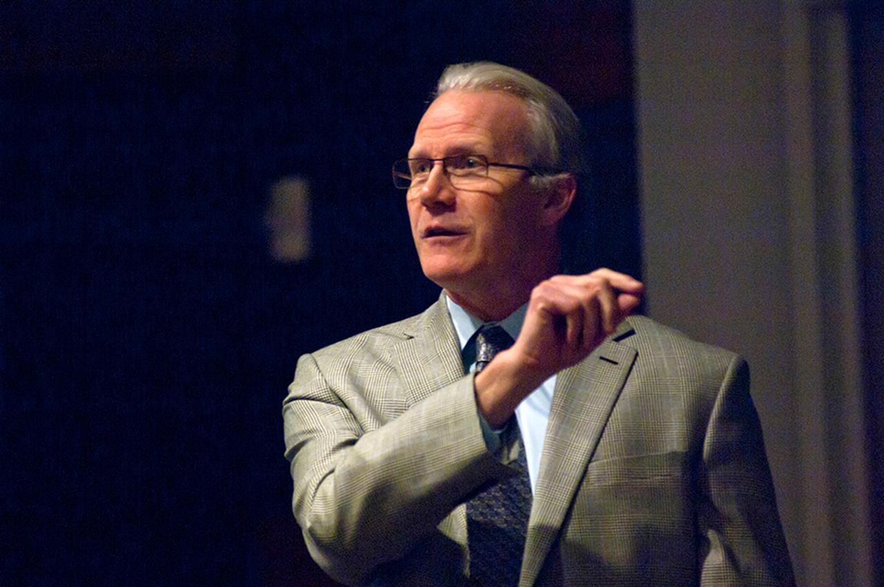 Randy Bryant speaks before an out-of-shot crowd. He has gray hair and wears a tan suit coat with a light blue line throughout and a blue shirt. 