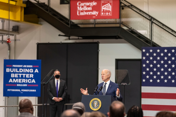 President Joe Biden wears a blue suit and stands behind a podium, addressing a crowd in an industrial environment. A Building a Better America sign is on his left, and the American flag is visible behind him to the right.