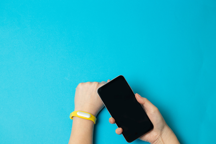 Two human forearms and hands rest on a turquoise table. The left wrist sports a yellow fitness tracker. The right hand holds a smartphone.