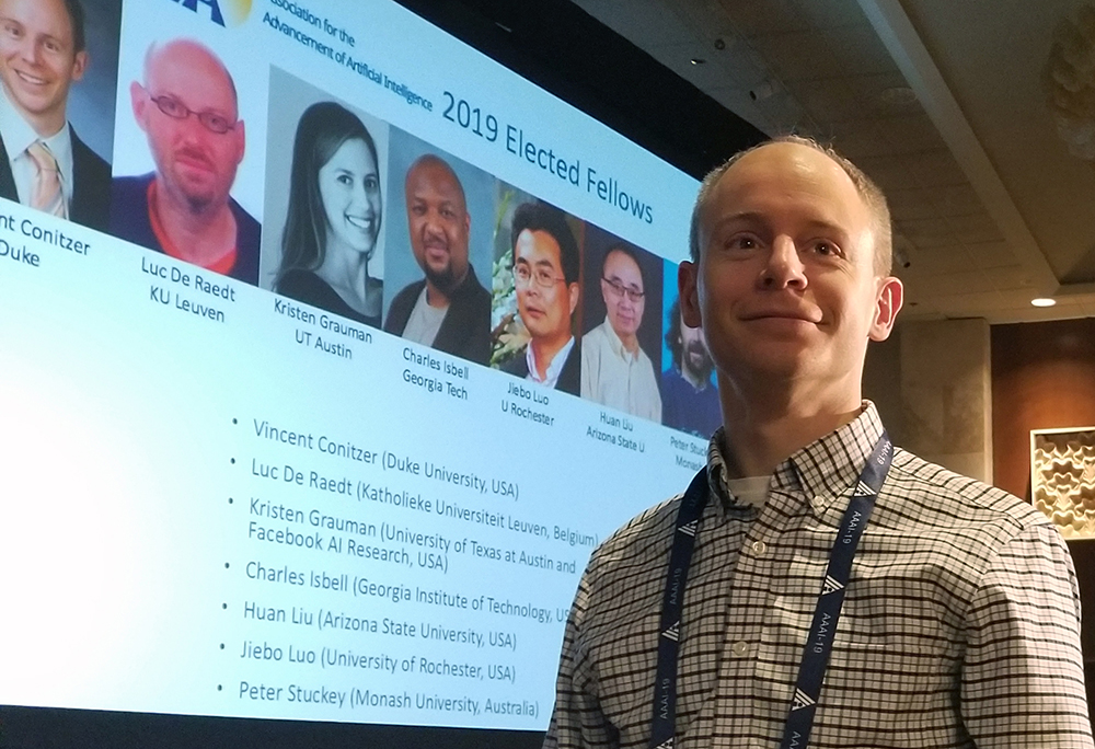 Portrait of Vince Conitzer, on the right, standing in front of a screen at a presentation screen at a conference.